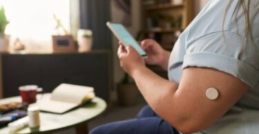 Wearable glucose sensor on an upper arm paired with a smartphone app, illustrating dual glucose-ketone sensor technology