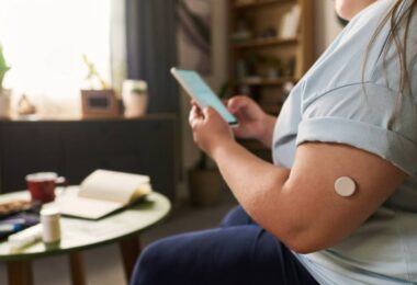 Wearable glucose sensor on an upper arm paired with a smartphone app, illustrating dual glucose-ketone sensor technology