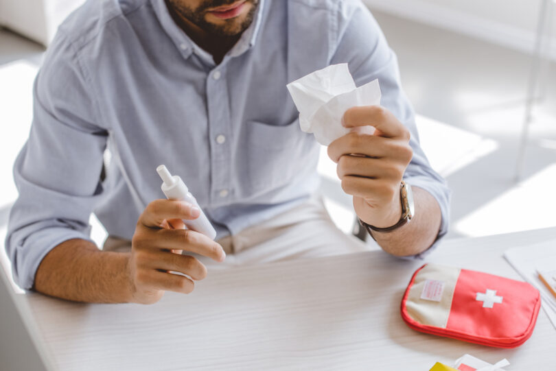 Cropped view of a person in an office holding a nasal spray and a tissue.